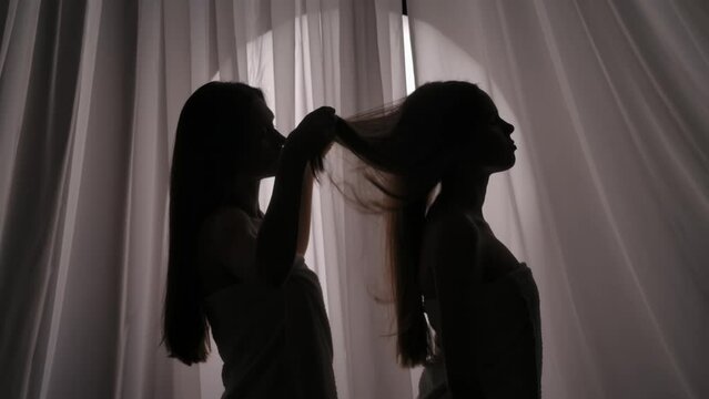 Medium side view video of two sisters, young women's silhouettes wrapped in a towel. One of them is stroking, touching the other one's hair in a muffled light.