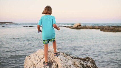 A five-year-old boy with long, light hair agilely leaps along the rocky seaside, exploring every corner.