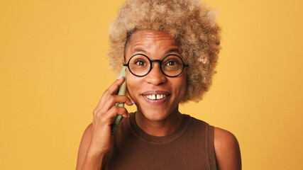 Close up, smiling girl in glasses, wearing brown top talking on mobile phone isolated on orange background