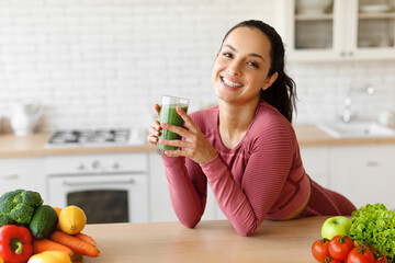 Arabic lady in sportswear holds glass with smoothie at kitchen