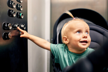 Tiny cute baby sitting in a baby stroller presses the floor button in the elevator. A little blonde...