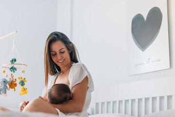 Mom breastfeeding her newborn baby in her arms in bed while smiling, in the background is a cradle...
