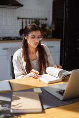 Woman in eyeglasses reading book near laptop and sticky notes on table at home, online education concept 