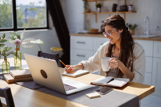 Smiling woman writing on notebook and holding cup near devices in kitchen at home