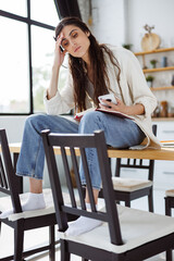 Tired woman holding smartphone and notebook while sitting on kitchen table at home