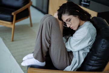 Tired woman in eyeglasses suffering from burnout while sitting on armchair at home