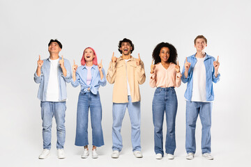 Group of young excited diverse friends looking and pointing fingers up at copy space standing over white background