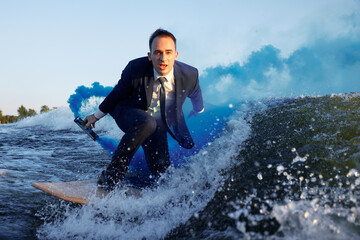 Young man in classic suit with blue smoke bomb in his hand rides wakeboard on a lake near city....