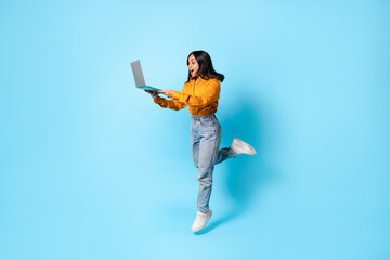 Excited indian lady holding laptop while jumping on blue background