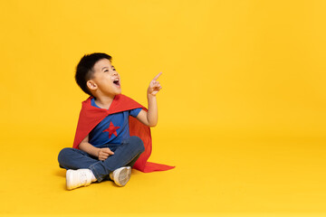 Superhero Asian boy with red cape sitting on floor and pointing to copy space isolated yellow...