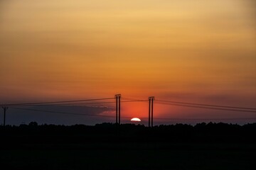 setting sun among the clouds in the red and orange sky, with electricity pylons in the middle and the forest line in the dark