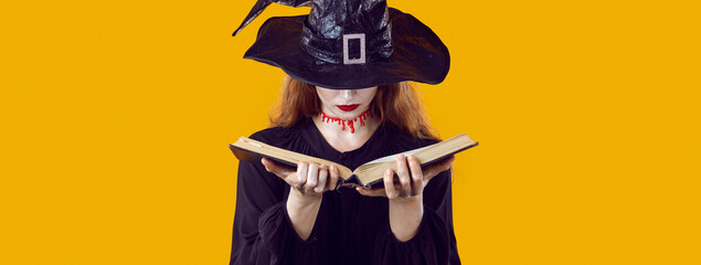 Evil witch reading book of spells. Beautiful young woman dressed as witch with bloody cut on her neck hiding her face under hat and looking through her witchcraft manual. Halloween and magic concept