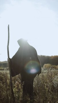 Amazing image of a witch. A woman with a stick in her hands walks forward across a weedy field. The witch is dressed in old black rags with a hood on her head. The person's face is hidden. Vertical