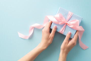 Celebratory scene: First person top view of my hands cradle a pastel blue gift box adorned with polka dots and a dainty pink ribbon bow. Against matching pastel blue backdrop, canvas for your message