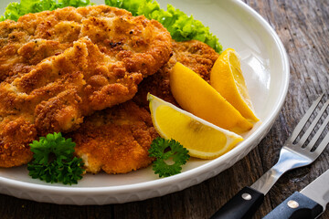 Crispy breaded seared chicken cutlet with lemon slices and fresh vegetables on wooden table
