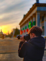 A teenage boy in a jacket takes a photo with a camera. Behind him is a blurred evening background in perspective