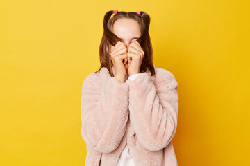 Shy brown haired girl wearing pink fur coat with ponytails isolated over yellow background covering her eyes with hair hiding her face.