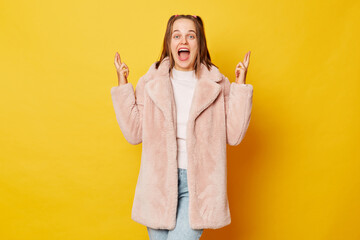 Pretty brown haired girl wearing pink fur coat with ponytails isolated over yellow background stands with crossed fingers warm smile emanating strong belief in positive outcomes - obrazy, fototapety, plakaty