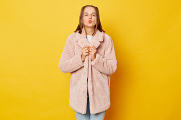 Yes I gained my goals. Joyful smiling brown haired girl wearing pink fur coat with ponytails shaking clenched fists celebrating win or success enjoying victory isolated over yellow background