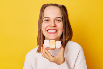 Joyful young girl with ponytails in jumper eating marshmallow at birthday party enjoying tasty...