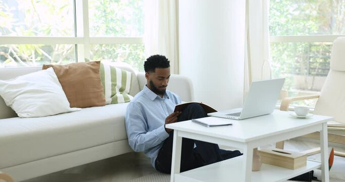 Handsome African man listen webinar, take part in webcast take notes in notebook seated on floor in cozy living room, modern tech usage to gain knowledge, studying, learn program, receive new skills