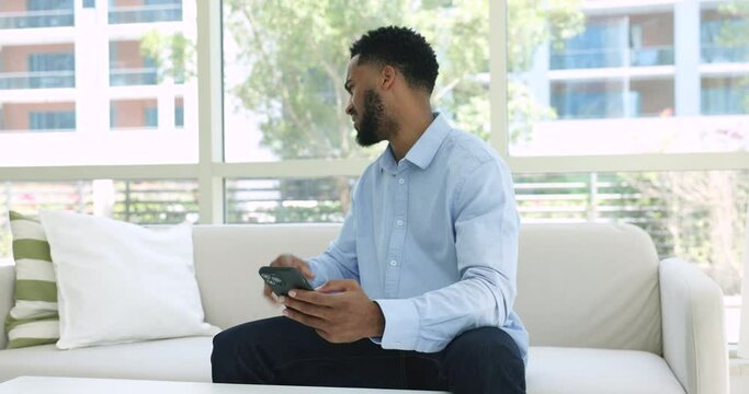 African man sit on sofa with cellphone freaking out, nervous reading bad news in a message feels frustrated get awful sms, personal or business concerns, gadget problems, unexpected badly information