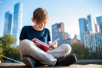 happy teenager reading a book in a metropolis park, against the backdrop of skyscrapers on a sunny day. Cute little boy reading a book sitting on a bench in the city