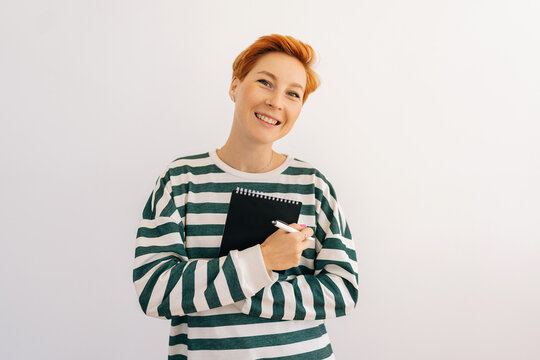 Studio portrait of cheerful young woman holding in hands closed paper notebook and pen, smiling looking at camera, standing on white isolated background. Cute female student making notes in copybook.