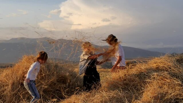 three happy children throw straw at each other having fun and playfully laughing and having fun in a rural field in the highlands at sunset. Brother and two sisters having fun outdoors