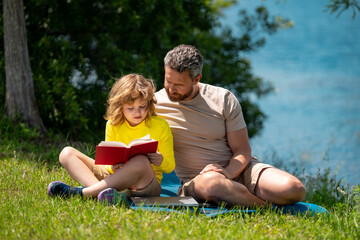 Naklejka na ściany i meble Father reading book with son in park, preparing school homework together, parenting. Summer lifestyle. Parenting and childhood concept. Little boy learning with father in outdoors garden.