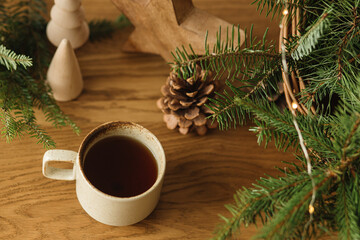 Winter holidays hygge. Stylish cup of warm tea, fir branches in basket, wooden trees and star, pine cones and lights on table. Modern christmas rustic eco friendly decor in scandinavian room