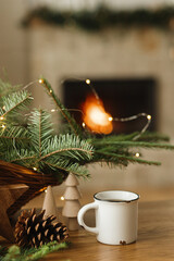 Stylish cup of warm tea, fir branches, wooden trees and star, pine cones on table against burning fireplace. Modern christmas rustic eco friendly decor and empty mug, scandinavian winter hygge