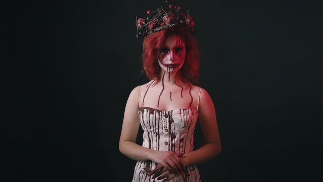 Face art woman with red hair and bloody face, in white bloody dress looking to camera on black background