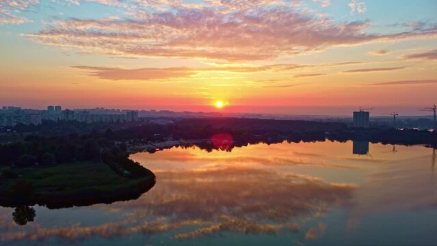 Aerial dolly pull out footage of sunrise sky on lake with clouds reflecting in calm water. Morning by the river in city park near residential district