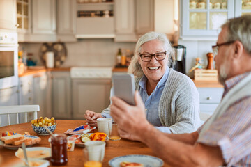 Senior Caucasian couple using a smartphone while having breakfast together in the kitchen in the morning