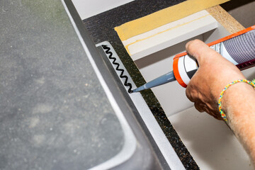 A man applies silicone caulk with a gun to the edge of a kitchen sink to install on the countertop.