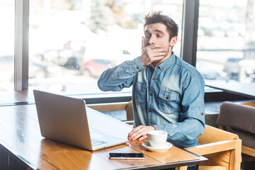 Portrait of handsome shocked scared young man freelancer in blue jeans shirt working on laptop,...