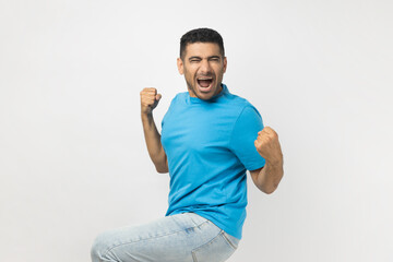 Portrait of unshaven man wearing blue T- shirt standing clenches fists, rejoices his victory and...