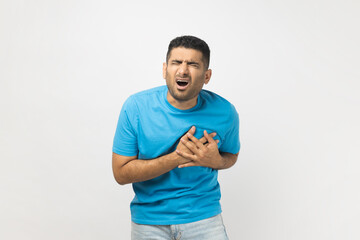 Portrait of stressed sick unshaven man wearing blue T- shirt standing with hands on chest, having heart attack, screaming from pain. Indoor studio shot isolated on gray background.