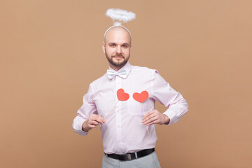 Portrait of cute friendly bald bearded man with nimb over head, holding little red hearts in front of his chest, wearing light pink shirt and bow tie. Indoor studio shot isolated on brown background.