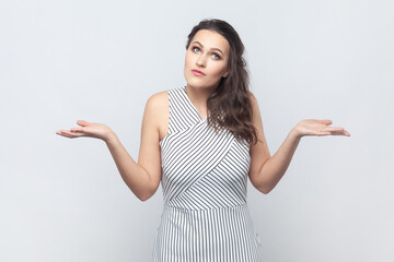 Portrait of brunette woman spreads palms, shrugs shoulders with perplexed expression, being indecisive, has no idea what happened, wearing striped dress. Indoor studio shot isolated on gray background - obrazy, fototapety, plakaty