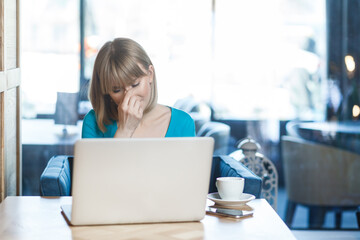 Portrait of sad despair upset young woman with blonde hair in blue shirt working on laptop, crying, hiding her face, having problems with her work. Indoor shot in cafe with big window on background.