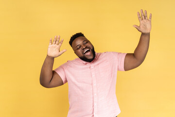Portrait of excited positive man with beard wearing pink shirt dancing, raising hands up and having...
