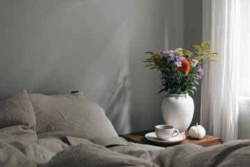 Elegant moody bedroom interior. Cup of coffee, pumpkin on retro wooden bedside table. White ceramic...