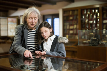 Friendly aged female tutor helping preteen girl exploring antique showpieces in local history museum..
