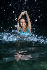 Obraz premium A beautiful and curly young woman in a blue swimsuit in the sea at night splashing water everywhere