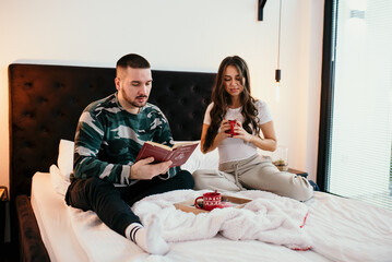 Young couple relaxing in bed reading a book and drinking coffee