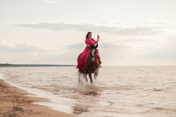 Beautiful young female in a bright pink dress riding a horse on a beach