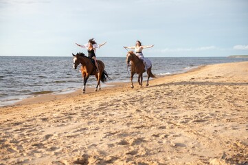 Two young female friends riding horses together on a sunny beach