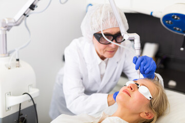 Naklejka premium Female client receiving face skin laser resurfacing procedure from professional beautician on modern equipment in clinic of aesthetic cosmetology